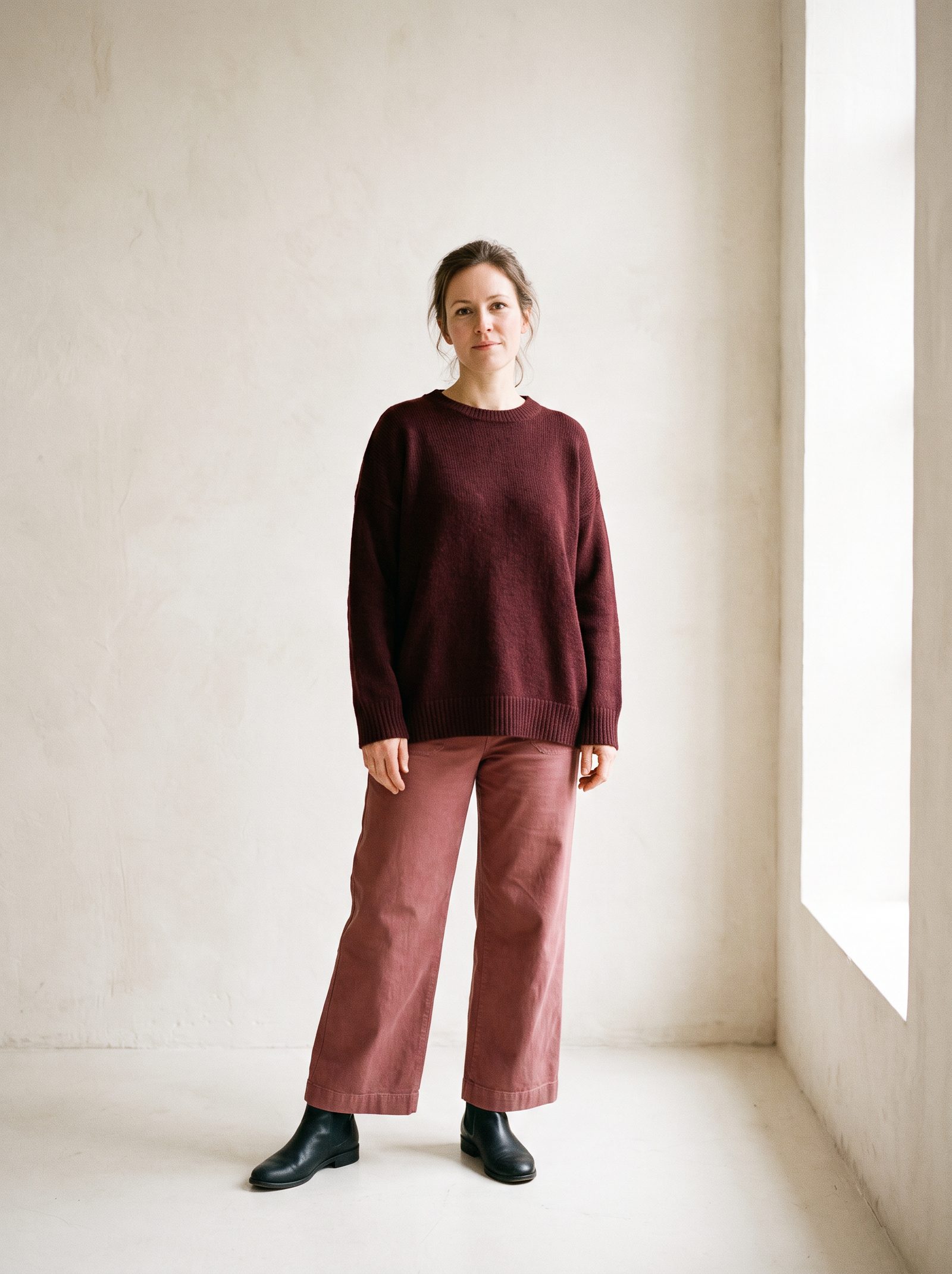 A full-body reference photo of a woman standing against a plain wall in casual clothes